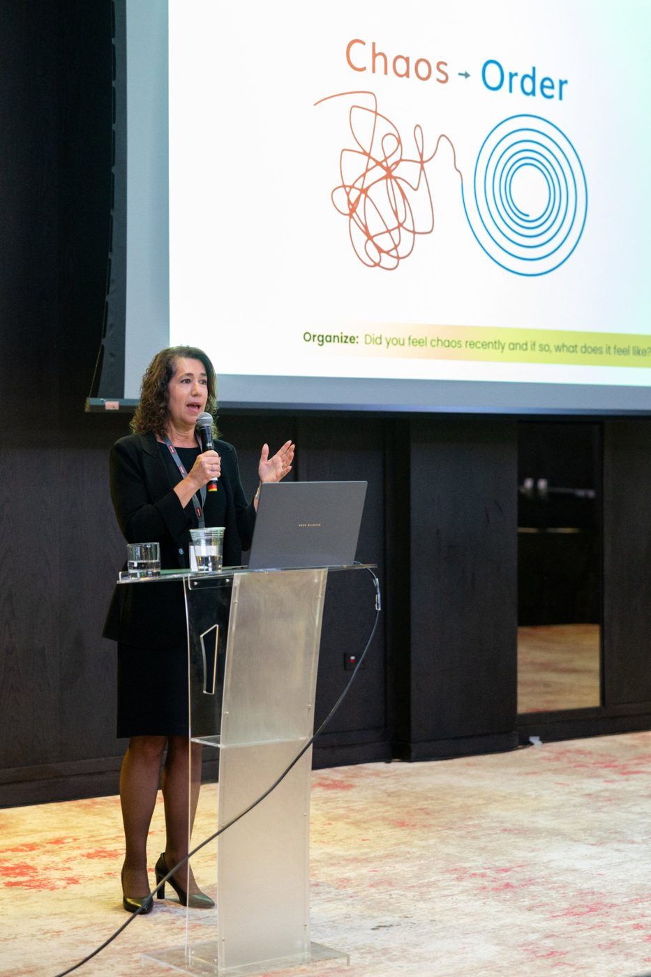 Sara Curwen speaking at a podium in front of a slide titled Chaos to Order